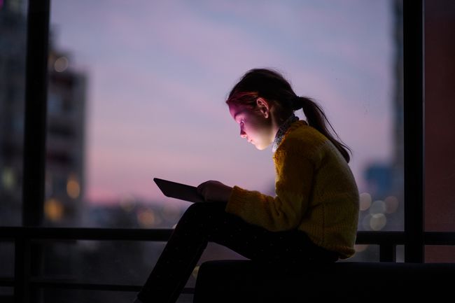 Lapsi istuu ikkunan edessä auringonlaskun aikaan suuren kaupungin yllä ja keskittyy tablettitietokoneeseen.